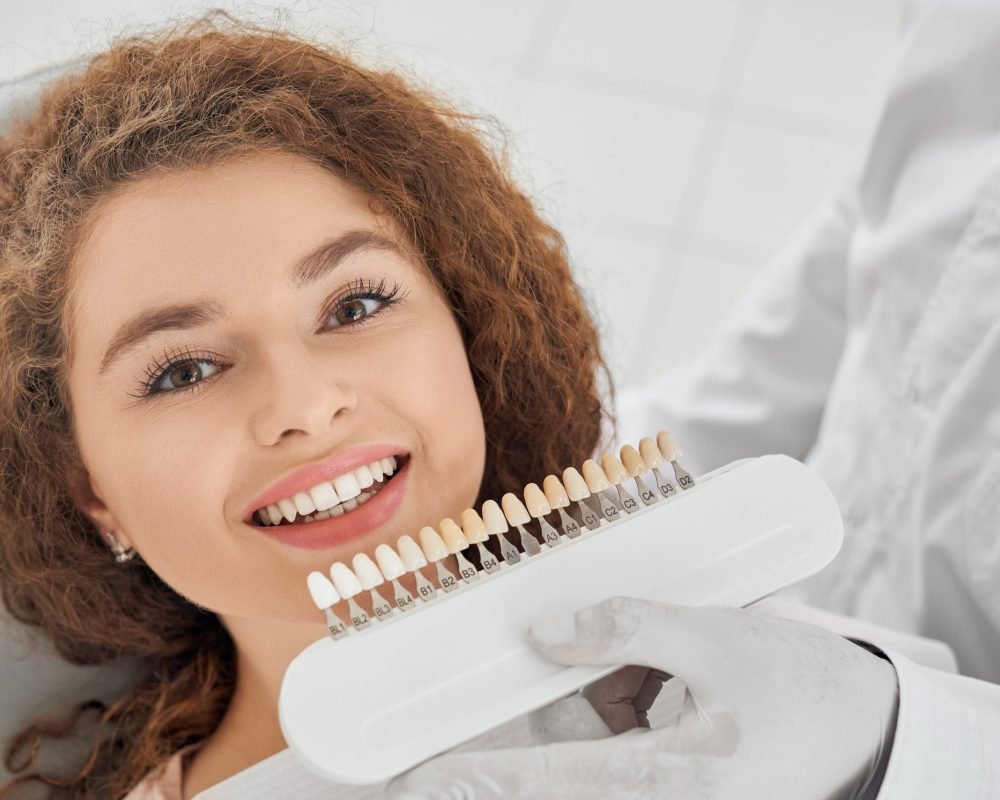 Woman smiling while male dentist keeping teeth color range Closeup of beautiful young woman lying on dental chair, looking at camera and smiling while male dentist keeping in hands teeth color range. Girl doing whitening procedure in dental office.