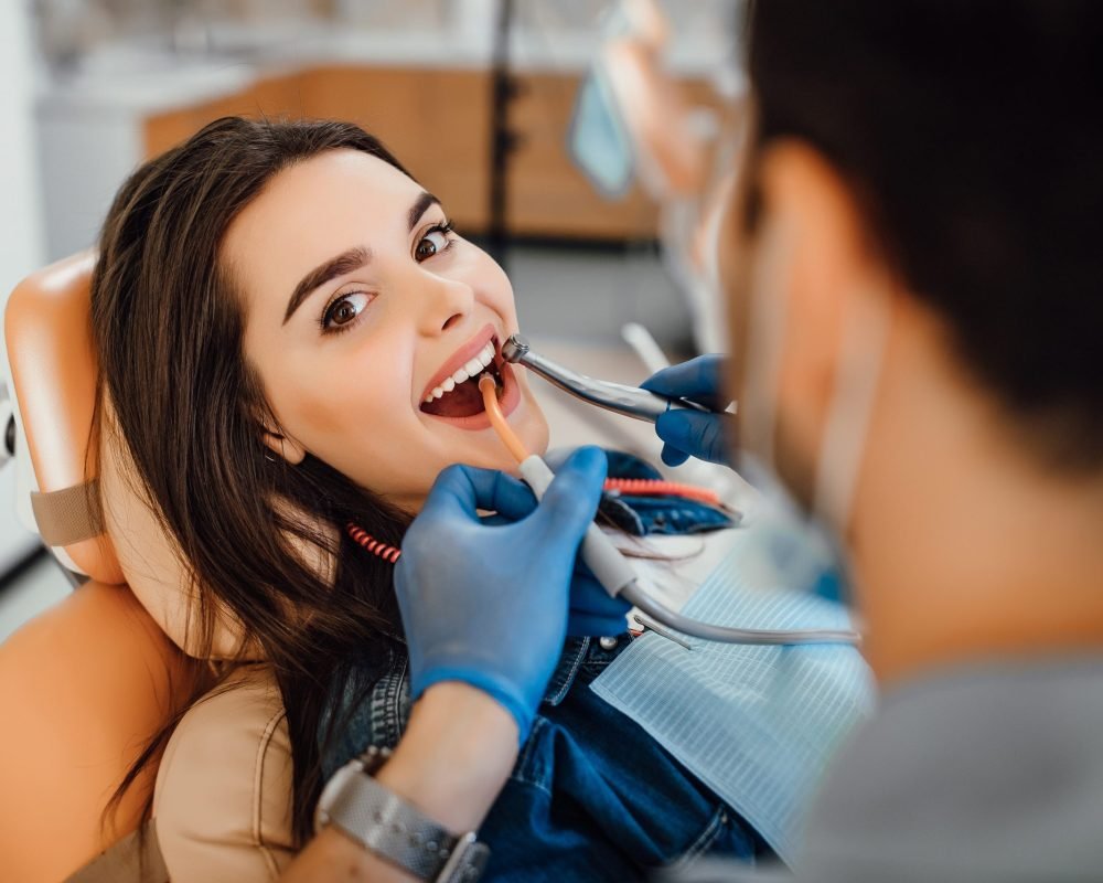 young-female-patient-visiting-dentist-office Young female patient visiting dentist office. Beautiful woman with healthy straight white teeth sitting at dental chair with open mouth during oral checkup while doctor working at teeth.